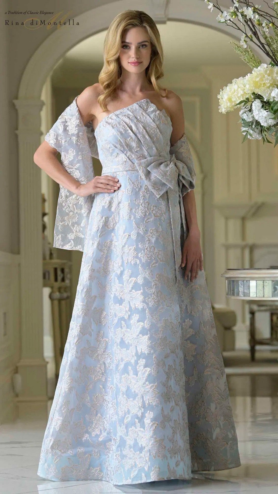 A woman wears the Long Formal Dress: Rina Di Montella RD3427 in blue, featuring a strapless jacquard design with silver floral patterns, an off-shoulder asymmetrical neckline, waist bow, and flowing A-line skirt.